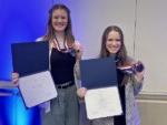 2 smiling students holding award medals and certificates
